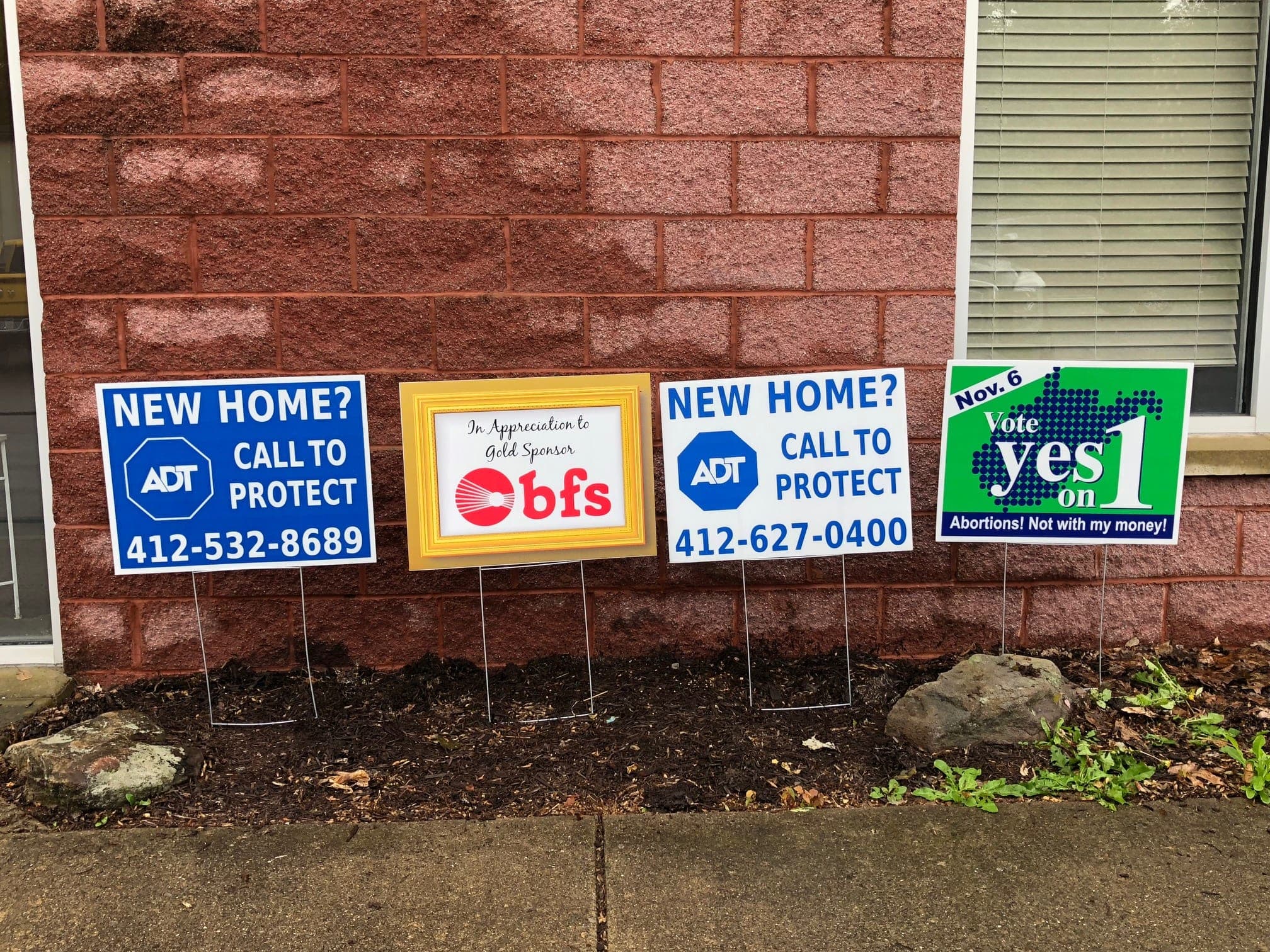 Row of printed yard signs, including ADT and BFS, staked in a flower bed